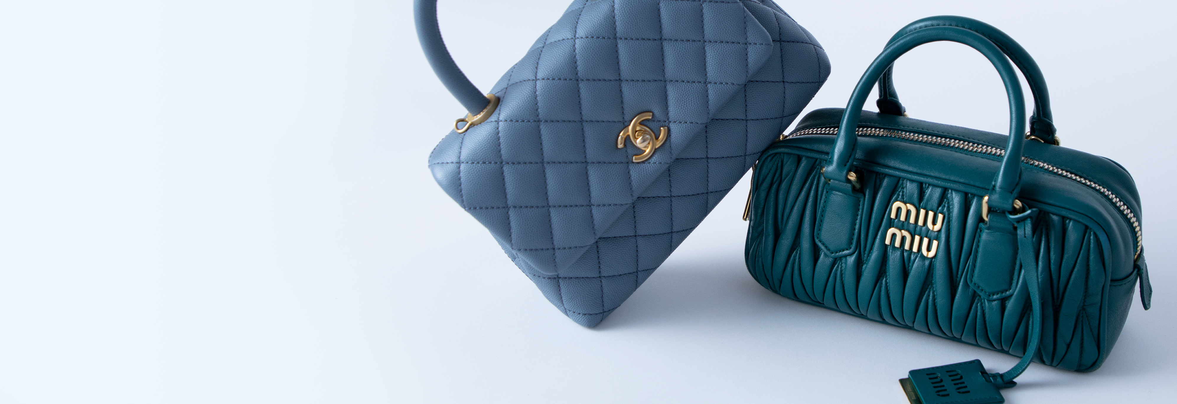 Light blue Chanel handbag leaning against a teal Miu Miu handbag against a sky blue background